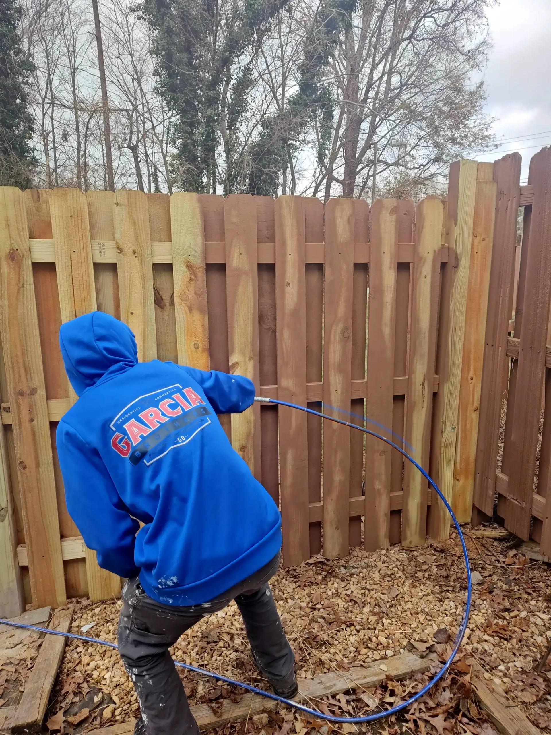 A man in blue jacket spaying the wood fence with brown paint.